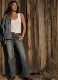 Fashion editorial: woman in kick-boot denim, white tank, concho belt, and boots against a barn wall at golden hour