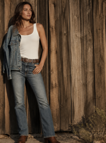 Fashion editorial: woman in kick-boot denim, white tank, concho belt, and boots against a barn wall at golden hour