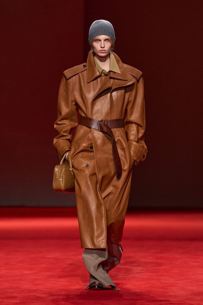Bottega Veneta Fall 2026 look 16: a long cognac leather trench coat cinched with a wide brown leather belt