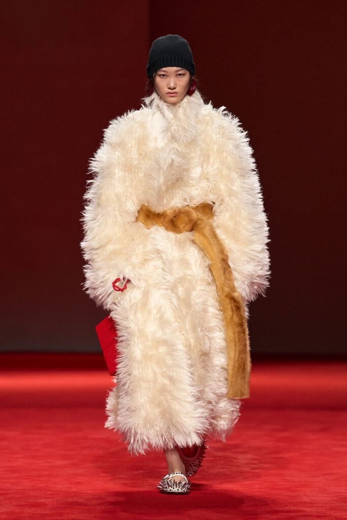 Bottega Veneta Fall 2026 look 40: a long cream shaggy coat tied with a wide camel belt, worn with a black beanie