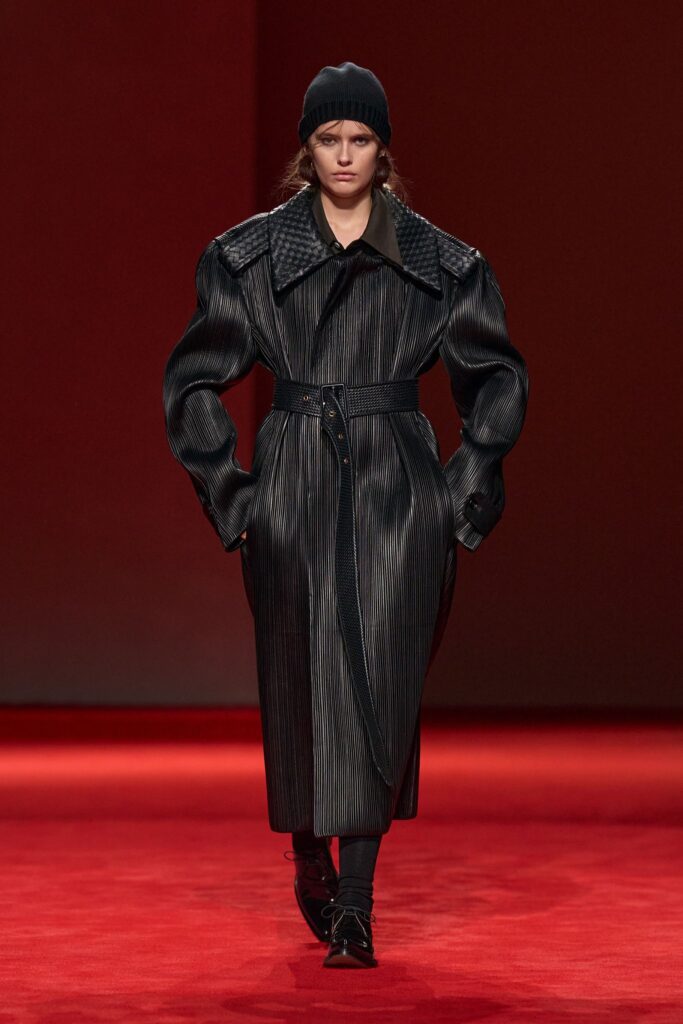 Bottega Veneta Fall 2026 look 55: a long black belted coat with a tightly ridged surface and a woven collar, worn with black boots