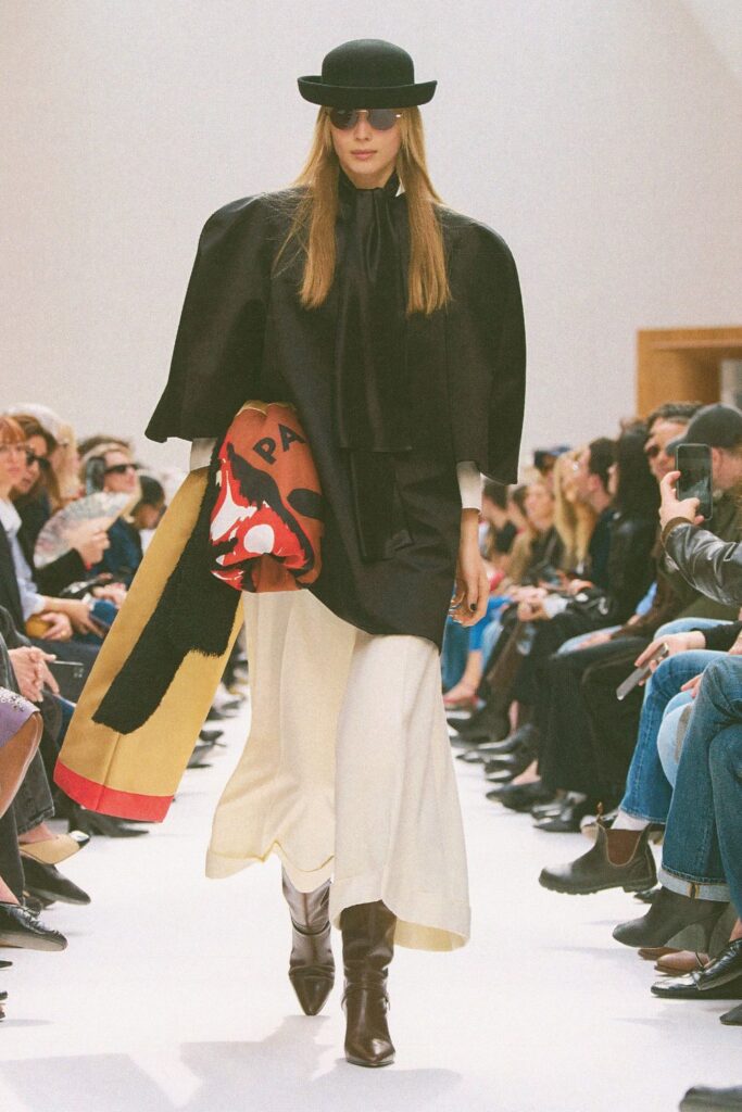 Celine Fall 2026 look 11: dark brown pointed-toe boots under a cream midi skirt, with a black jacket, black bowler hat and a printed scarf