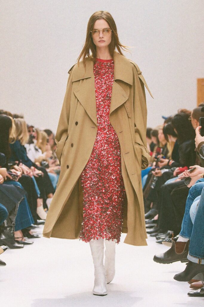 Celine Fall 2026 look 32: white pointed mid-calf boots with a red sequined midi dress and an open camel trench coat