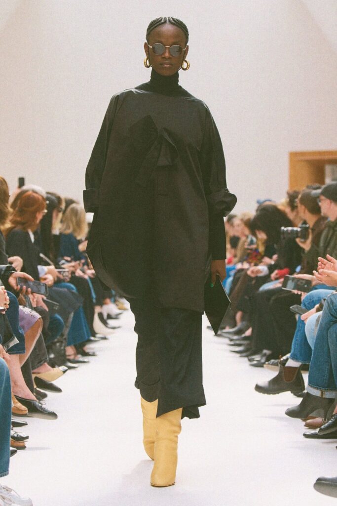 Celine Fall 2026 look 43: pale yellow mid-calf boots worn with an all-black look of a draped coat and long skirt