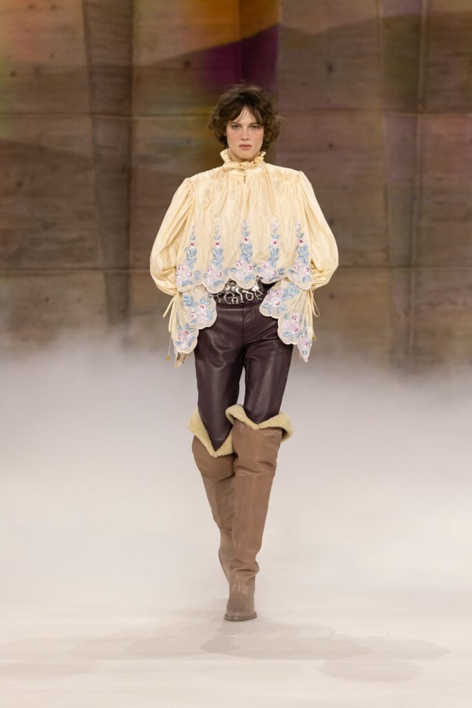 Chloé Fall 2026 look 36: cream embroidered high-neck blouse, logo buckle belt and brown leather trousers tucked into tall tan boots