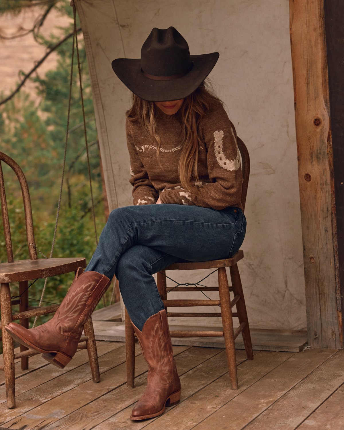 Woman in a brown Western-motif sweater, jeans, dark cowboy hat and brown cowboy boots seated on a wooden porch