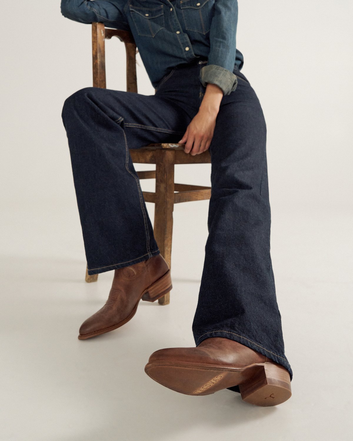 Brown leather cowgirl boots with a low stacked heel under dark flare jeans and a denim shirt