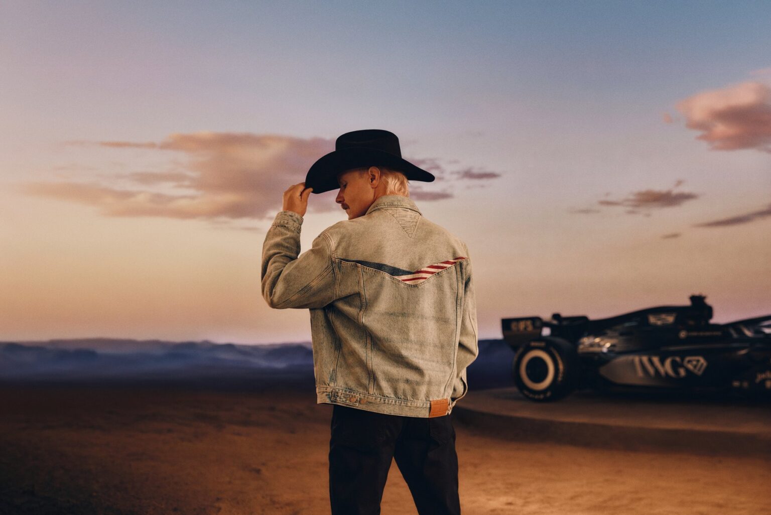 Back view of a light-wash denim trucker jacket with flag-stripe detail and a black cowboy hat at dusk beside a Cadillac F1 car