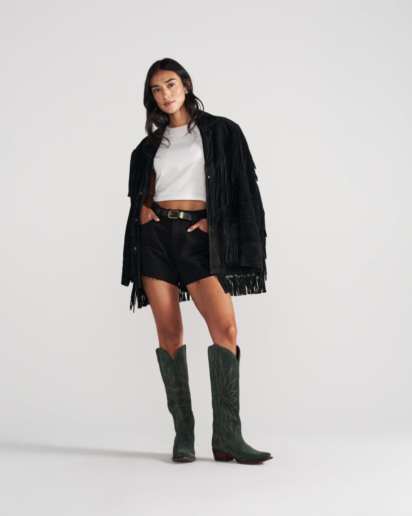 Black suede fringe jacket over a white crop tee with black shorts and tall green cowgirl boots